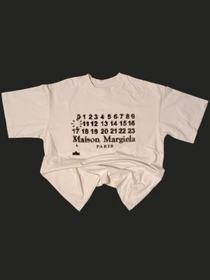 vit t-shirt med print - Vit t-shirt med ikoniskt svart print framtill som visar siffror och logga. Modellen är kortärmad med rund hals och har en loose passform. Materialet är mjuk bomull som känns skönt mot huden. Perfekt för dig som gillar statement-plagg och designer vibes.