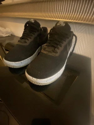 Svarta Nike Air Force 1 sneakers - Säljer ett par klassiska Nike Air Force 1 sneakers i svart med vit sula. Skorna har snörning och är tillverkade i textilmaterial med rund tå och platt sula. Perfekta för dig som gillar streetstyle och vill ha en tidlös look.