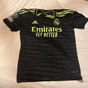 Real Madrid Vini Jr. matchtröja XS - Säljer en svart och neongul Real Madrid matchtröja från Adidas i storlek XS. Tröjan har Vini Jr. och nummer 20 på ryggen, samt Champions League-märke på ärmen. Tillverkad i polyester med korta ärmar och sportig design.