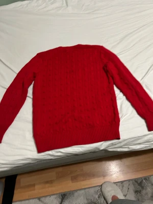Röd stickad tröja från Polo Ralph Lauren - Riktigt snygg röd kabelstickad tröja från Polo Ralph Lauren med klassisk rund halsringning och broderad marinblå logga på bröstet. Tröjan har långa ärmar och breda ribbade muddar vid ärmslut och nederkant. Perfekt för dig som vill ha en stilren och tidlös look. Priset kan sänkas vid snabb köp