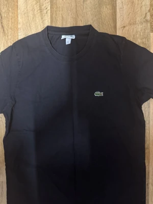 Svart t-shirt från Lacoste - Svart t-shirt från Lacoste med klassisk rund hals och det ikoniska gröna krokodilmärket broderat på bröstet. Tillverkad i mjuk bomull och har en enkel, stilren design som passar perfekt till jeans eller shorts.