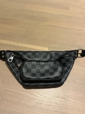 En ruttig axelväska  - Snygg axelväska med klassiskt rutigt mönster i svart och grått. Väskan har flera fack med dragkedja och justerbar rem. Materialet är skinn och detaljerna i silverfärgad metall ger en lyxig vibe. Perfekt för att bära det viktigaste med stil.