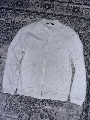 Grå bomberjacka från Zara - Snygg grå bomberjacka från Zara med stilren design. Jackan har ribbad krage, muddar och nederkant, samt två sidofickor. Materialet är mjukt och känns som bomull, med hel dragkedja framtill. Perfekt för dig som gillar enkel och clean stil.