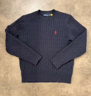 Ralph Lauren stickad  - Kabelstickad mörkblå tröja från Ralph Lauren med klassisk rund hals och röd broderad logga på bröstet. Långärmad modell i mjuk ull, perfekt nu under lite kyligare dagar!