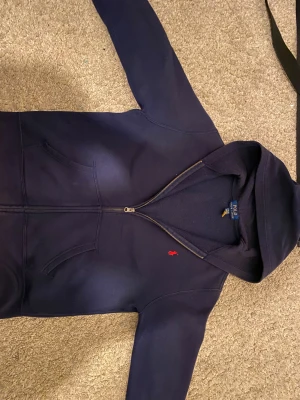 Mörkblå hoodie från Polo Ralph Lauren - Snygg mörkblå hoodie från Polo Ralph Lauren med dragkedja och huva. Tröjan har ett rött broderat Polo-logo på bröstet och två praktiska fickor framtill. Tillverkad i mjuk bomull, perfekt för chill dagar. Klassisk och stilren design som alltid funkar. Köpte den i NK i stockholm 