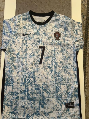 Portugal Ronaldo fotbollströja - Säljer en Portugal fotbollströja från Nike med Ronaldo och nummer 7 på ryggen. Tröjan har ett unikt blått och vitt mönster, svart krage och korta ärmar. Materialet är lätt och andas, perfekt för match eller träning. Landslagsemblem på bröstet och Nike-logga.