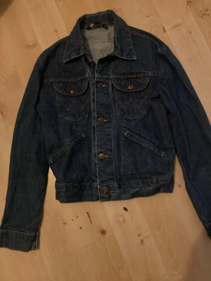 Mörkblå jeansjacka från Wrangler - Snygg mörkblå jeansjacka från Wrangler med klassisk design, bruna metallknappar och två bröstfickor med W-söm. Jackan har lång ärm, krage och raka linjer. Perfekt för dig som gillar tidlös denim och vill ha en cool look.