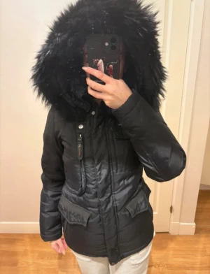 Svart pufferjacka med pälsluva - Säljer en svart pufferjacka från Hollies med stor fluffig päls på luvan. Jackan har dragkedja och knappar framtill, flera fickor med lock och en badge på ärmen. Perfekt för kalla dagar och har en snygg, figurnära passform. Använd endast en vinter