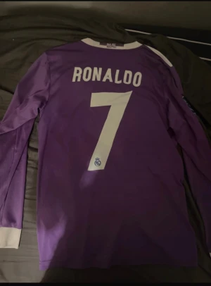 Real Madrid Ronaldo långärmad tröja - Säljer en officiell Real Madrid långärmad fotbollströja från Champions League-finalen 2017 med Ronaldo 7 på ryggen. Tröjan är lila med vita detaljer, Adidas-logga, Fly Emirates sponsortryck och flera mästerskapsmärken. Materialet är lätt och andas, Climacool-teknologi.