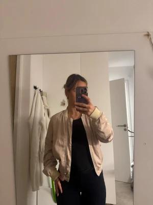 Beige bomberjacka med dragkedja - Säljer en beige bomberjacka med ribbade muddar i ljusare ton och dragkedja framtill. Jackan har en klassisk, loose passform och är tillverkad i ett syntetmaterial som ger en lätt och skön känsla. Perfekt att slänga på sig för en chill och stilren look.