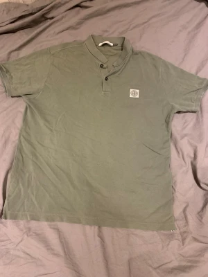 Grön pikétröja från Stone Island - Säljer en stilren grön pikétröja från Stone Island med klassisk krage och två knappar framtill. Tröjan har korta ärmar och en snygg broderad logga på bröstet. Tröjan är perfekt för dig som vill se elegant och stilig ut.