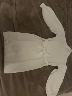 Vit stickad klänning med knytband - Superfin vit stickad klänning med långärmade puffärmar och ett snyggt knytband i midjan. Klänningen har ribbade detaljer vid halsen och nederkanten, vilket ger en extra mysig känsla. Perfekt för dig som gillar stilrena och bekväma plagg.