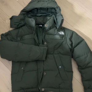 Grön pufferjacka från The North Face - Säljer en mörkgrön pufferjacka från The North Face i storlek XS. Jackan har huva, dragkedja och knappar framtill samt två stora fickor. Perfekt för kalla dagar och har en clean look med logga på bröstet och ryggen.