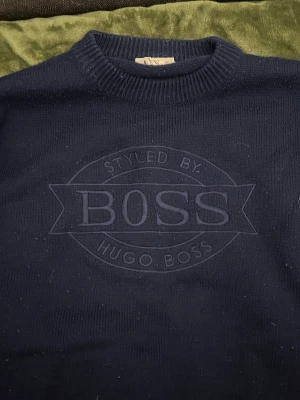 Mörkblå stickad tröja från Hugo Boss - Säljer en mörkblå stickad tröja från Hugo Boss Sports med stort broderat BOSS-logo på bröstet. Tröjan har rund halsringning, ribbade muddar och en relaxed passform. Perfekt för dig som gillar klassisk streetwear med premiumkänsla. Den är nopprig därför priset är lägre