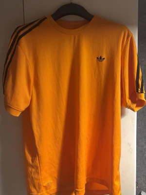 Orange Adidas t-shirt med svarta ränder - Säljer en fräsch orange t-shirt från Adidas med klassiska svarta ränder längs axlarna och det ikoniska Adidas-loggan på bröstet. T-shirten har rund hals och korta ärmar, tillverkad i mjuk bomull som är skön mot huden. Perfekt för dig som gillar streetwear och sportig stil.