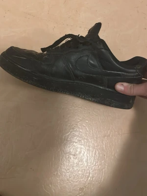 Svarta Nike Air Force 1 skor - Svarta Nike Air Force 1 sneakers med klassisk låg siluett och rund tå. Skorna har snörning och är tillverkade i skinn med en platt sula. Perfekt för dig som gillar streetwear och vill ha en ikonisk sneaker i garderoben.