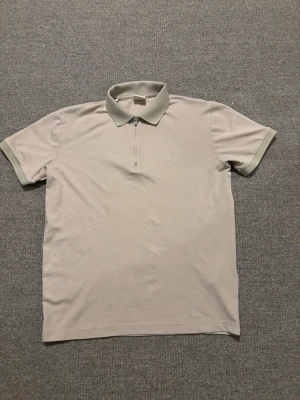 Beige polotopp från Selected Homme - Säljer en stilren beige polotopp från Selected Homme med korta ärmar och krage i ett luftigt matrial som är skön att ha på sommaren. Toppen har en diskret dragkedja framtill och ribbade detaljer vid ärmslut och krage. Perfekt för dig som gillar minimalistisk design.