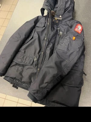 Svart Parajumpers parkajacka med huva - Svart parkajacka från Parajumpers med stor huva, flera fickor och snygga detaljer som patch på ärmen och orange inslag. Jackan har quiltad insida och mjuk fuskpäls vid nacken. Perfekt för kalla dagar och har dragkedja samt knappar framtill.