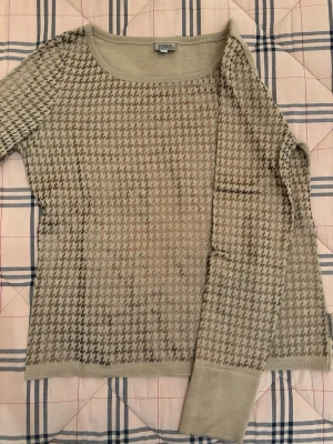 Beige rutig tröja från Ferre Jeans - Snygg beige tröja från Gianfranco Ferre Jeans i storlek M. Tröjan har ett klassiskt rutigt mönster i bruna toner och är stickad med rund halsringning och långa ärmar. Perfekt för dig som gillar stilrena detaljer och mjuka material.