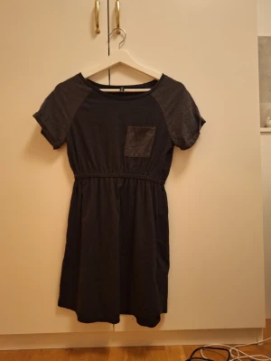 Svart vintage kortklänning  - Svart kortklänning med rund hals, korta ärmar och en liten bröstficka. Klänningen har resår i midjan för en snygg passform och är gjord i mjuk bomull. Perfekt för dig som gillar enkel och stilren design.