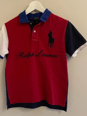 Röd och marinblå Polo Ralph Lauren piké - Snygg piké från Polo Ralph Lauren i rött och marinblått med vit detalj på ena ärmen. Klassisk pikékrage i blått, broderad logga och text på bröstet. Kortärmad och tillverkad i mjuk bomull. Perfekt för dig som gillar sportig och stilren look. Säljs då jag har vuxit ur den. Den är i storlek 10-12 år