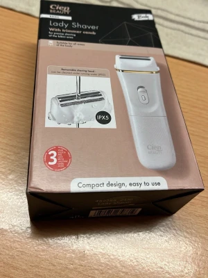 Cien Beauty Lady Shaver - Elektrisk lady shaver i vit plast med gulddetalj, designad för att raka och trimma kroppshår. Kommer med avtagbart rakhuvud, trimmerkam och rengöringsborste. IPX5-klassad för enkel rengöring under vatten. Kompakt och lättanvänd.