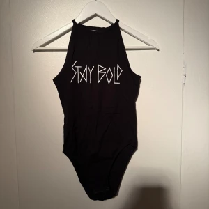 Svart body - Svart bodysuit med hög hals och ärmlös design. Framsidan har ett vitt tryck med texten 'STAY BOLD' i grafisk stil. Storlek s från hm