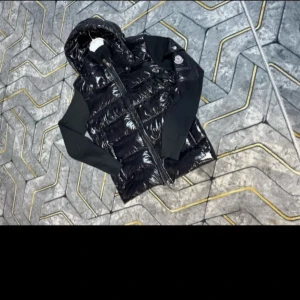 Svart puffer kardigan jacka med huva  från Moncler - Snygg svart puffer kardigan jacka  från Moncler med glansig yta och huva. Jackan  har quiltad design, dragkedja framtill och ribbade svarta ärmar.med NFC Patch QR kod  Moncler-logga på axeln ger en exklusiv touch. Perfekt för dig som vill ha en cool och sportig look.