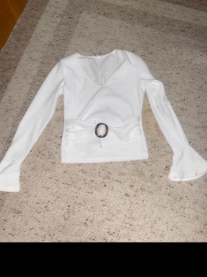 Vit omlottopp från Chiara Forthi - Säljer en vit omlottopp från Chiara Forthi med v-ringning och långa trumpetärmar. Toppen har ett brett midjeband med dekorativt spänne framtill och är ribbad i materialet för extra struktur. Perfekt för dig som gillar stilrena detaljer.liten smink fläck men går bort i tvätten 