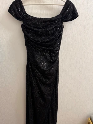 Svart glittrig långklänning - Svart långklänning med glittriga paljetter över hela tyget. Klänningen har offshoulder-ärmar, draperad framsida och en slits nedtill. Stängs med dragkedja i ryggen. 