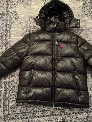 Svart pufferjacka från Polo Ralph Lauren - Säljer en svart pufferjacka från Polo Ralph Lauren med röd logga på bröstet. Jackan har huva med tryckknappar, tre dragkedjeförsedda fickor och en glansig finish. Perfekt för kalla dagar och har en riktigt schysst streetstil.