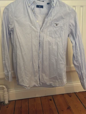 Ljusblå skjorta från GANT - Snygg ljusblå skjorta från GANT med klassisk button-down krage och bröstficka med broderad logga. Skjortan har långa ärmar och knappar framtill. Perfekt för dig som gillar stilrena och enkla plagg med en preppy vibe.