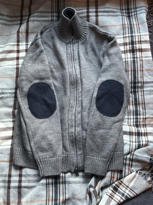 Grå kofta från Marithé + François Girbaud - Säljer en grå kofta med hög krage och hel dragkedja från Marithé + François Girbaud. Köpte den i barcelona. Tröjan har mörkblå armbågslappar, ribbade muddar och knappar som sitter under dragkedjan, som skapar en extremt snygg ”lager på lager” effekt. Perfekt för kyliga dagar, eller bara kalla sommarkvällar och riktigt snygg till jeans. Tröjan passar S och M beroende på hur man vill att den ska sitta. Hör av dig vid funderingar.