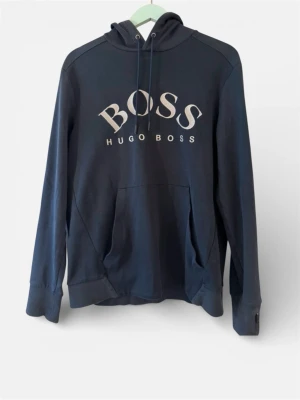 Hugo Boss blå hoodie, storlek M - Mörkblå hoodie från Hugo Boss med dragkedja vid ärmslut och luva med dragsko. Stor logotyp framtill.
