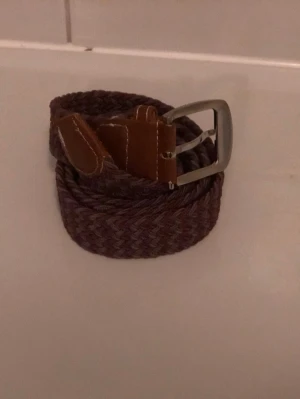 Lila och grå flätad bälte med läderdetaljer - Säljer ett brunt flätat bälte med snygga läderdetaljer och silverfärgat spänne. Bältet är cirka 110 cm långt och har en klassisk look som passar till jeans eller chinos. Perfekt accessoar för att lyfta din outfit. Köpt för 400kr