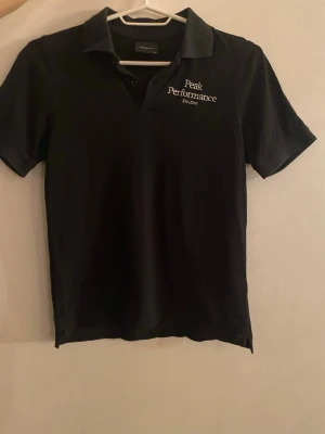 Svart Peak Performance polotopp - Svart polotopp från Peak Performance med klassisk krage och korta ärmar. Broderad vit logga på bröstet med texten 'Peak Performance Est. 1986'. Topp i mjuk bomull, perfekt för en clean och sportig look.Peak Performance Pikétröja Svart Est. 1986 Logotyp Kortärmad Krage Knappar Sportig Casual Kläder Herr Mode Stil Bekväm Hög kvalitet Bomull Pique Tyg Andningsaktiv Funktionell Golf Tennis Fritid Vardag Snygg Klassisk Tidlös Enkel Mångsidig Lätt Resa Semester Skandinavisk design Svensk design Outdoo