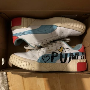 Puma sneakers med hjärtmönster - Säljer ett par vita Puma sneakers med ljusblå detaljer och röd häl. Skorna har coolt hjärtmönster och stor PUMA-logga på sidan. Klassisk snörning och platt sula. Perfekt för dig som gillar färg och streetstyle.
