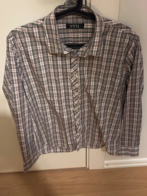 Rutig skjorta från STIL i storlek L - Säljer en klassisk rutig skjorta från STIL i storlek L (41-42). Skjortan har en vit bas med svarta och röda rutor, knappar framtill och långärmad design. Perfekt för dig som gillar en chill och stilren look. Materialet är mjuk bomull som känns skönt mot huden.