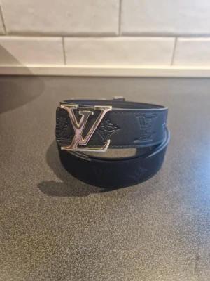 Svart Louis Vuitton bälte med LV-spänne - Säljer ett svart Louis Vuitton bälte med präglat LV-mönster längs hela remmen. Bältet har ett stort metallspänne med LV-logga i guld. Materialet är skinn och designen är klassisk med lyxig känsla. Perfekt för att lyfta din outfit.