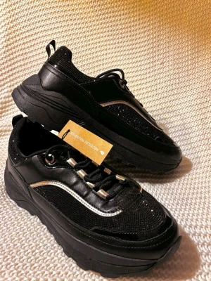 Svarta sneakers med silverdetaljer - Säljer ett par helt nya svarta sneakers med grov sula och snygga silverdetaljer vid snörningen och längs sidan. Ordinarie pris 449kr aldrig använda, lapp sitter kvar