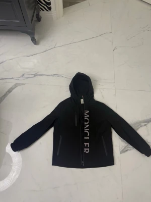 Svart vindjacka från Moncler - Svart vindjacka från Moncler med stor logotyp längs dragkedjan och klassisk patch på ärmen. Jackan har huva, två sidofickor med dragkedja och är tillverkad i polyester. Perfekt för dig som gillar stilrena och sportiga plagg.