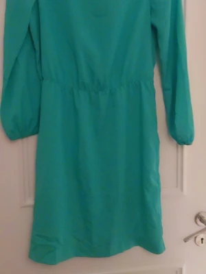 Grön  långärmad klänning från Åhléns  - Snygg grön klänning med lång ärm och rund hals. Modellen är enkel med resår i midjan som ger en skön passform. Materialet känns som polyester och klänningen har en slät yta utan mönster eller detaljer. Perfekt för dig som gillar färg och stilren design.