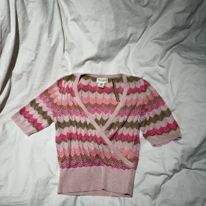 Rosa omlottkofta Holly & Whyte XS - Säljer en söt omlottkofta från Holly & Whyte i storlek XS. Koftan har ett zigzag-mönster i rosa, beige och olivgrönt, med ribbade kanter och korta ärmar. Materialet är stickat och känns mjukt mot huden. Perfekt för vår och sommar!
