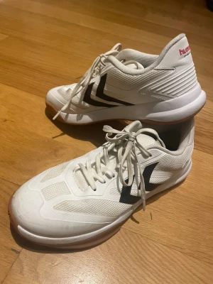 Vita sneakers från Hummel strl 39 - Säljer ett par vita Hummel sneakers med svart logga på sidan och gummisula i ljusbrun ton. Skorna har snörning och är tillverkade i syntetmaterial och mesh för bra ventilation. Perfekta för dig som gillar sportig stil och vill ha bekväma skor. Säljes pga felköp, storlek.