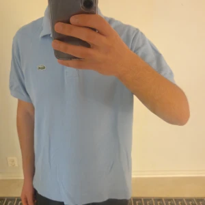 Ljusblå pikétröja från Lacoste - Säljer en klassisk ljusblå pikétröja från Lacoste i storlek L med den ikoniska krokodilloggan på bröstet. Tröjan har korta ärmar, krage och knappar framtill. Tillverkad i mjuk bomull som känns skön mot huden. Perfekt för dig som gillar stilrena och sportiga plagg.
