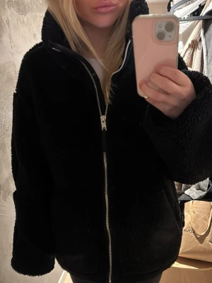 Svart teddyjacka med dragkedja - Mysig svart teddyjacka med hög krage och grov dragkedja framtill. Jackan har en oversized passform och är tillverkad i fluffigt teddy-material som håller dig varm. Perfekt för kalla dagar och enkel att styla till olika outfits.