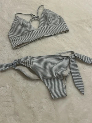 Randig bikini från H&M, storlek 34 - Säljer en fräsch bikini från H&M i storlek 34. Bikinin är vit med tunna blå ränder och har knytband på sidorna av underdelen samt korsade axelband på överdelen. Materialet är polyester, polyamid och elastan, vilket gör den mjuk och följsam. Lite missfärgning på underdelen, men inget man tänker på (se sista sidan)🫰🏼.