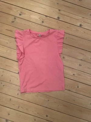 Rosa ribbad topp med volang från Pigalle - Säljer en ljusrosa ribbad topp från Pigalle i storlek S. Toppen har volangdetaljer vid axlarna och är ärmlös med rund halsringning. Materialet är mjukt och stretchigt, perfekt för vår och sommar. Supersöt att styla med jeans eller kjol!