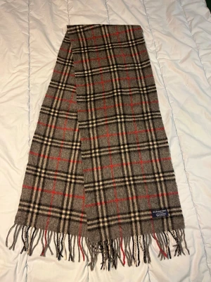 Grå rutig Burberry halsduk i kashmir - Säljer en klassisk grå halsduk från Burberry med rutigt mönster i svart, vitt och rött. Västen är tillverkad i 100% kashmir och har fransar längs nederkanten. Perfekt för dig som gillar tidlös design och mjuka material. N17