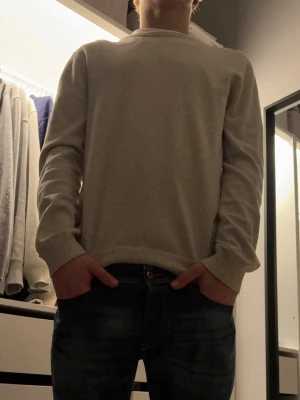 Stickad biege tröja från bläck - En stilren ljusgrå långärmad tröja från bläck med rund hals och ribbade muddar vid ärmslut och nederkant. Tröjan har en normal passform och är tillverkad i mjuk bomull, perfekt för chill dagar eller när du vill hålla det enkelt.